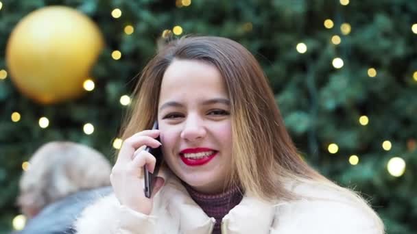 スマートフォンで話している若いブロンドの女の子は ぼやけたクリスマスツリーの背景 スローモーション — ストック動画