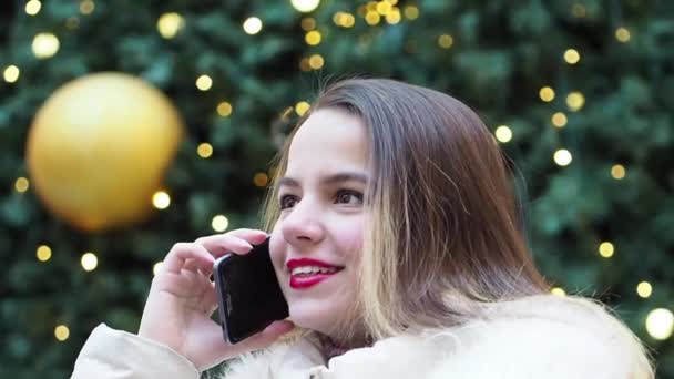 スマートフォンで話している若いブロンドの女の子は ぼやけたクリスマスツリーの背景 スローモーション — ストック動画