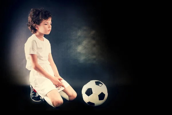 Sad and not happy little kid with football about lost football or soccer game. Dark background. Copy space.