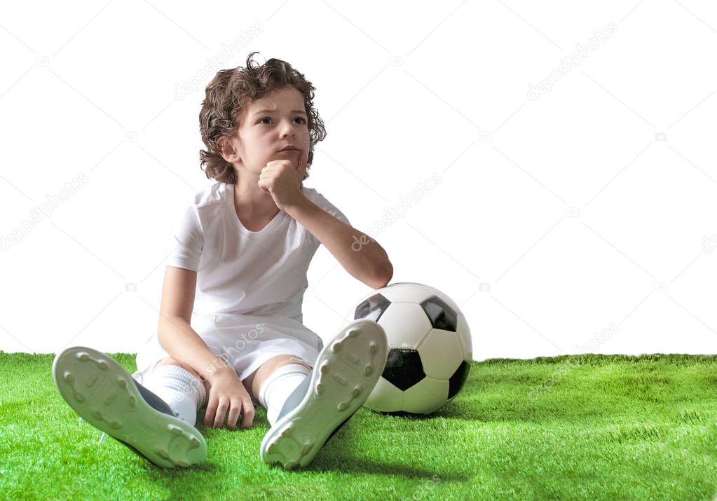 Puzzled little footballer sitting on the grass. Copy space