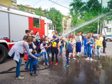 Moskova, Rusya - 07 Ağustos 2019: İtfaiye istasyonunda yangın hortumu deneyen ve su püskürten, bir itfaiyecinin yardım ettiği çocukların ve ebeveynlerin yan görünümü