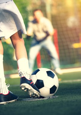 Babasıyla futbol sahasında oynayan küçük bir çocuk. Çocuk futbolcu sahada atış yapmaya hazırlanıyor. Futbol sahasında genç bir kaleci futbol kapısında duruyor..