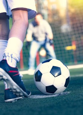 Babasıyla futbol sahasında oynayan küçük bir çocuk. Çocuk futbolcu sahada atış yapmaya hazırlanıyor. Futbol sahasında genç bir kaleci futbol kapısında duruyor..