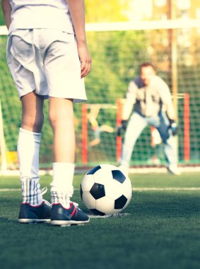 Babasıyla futbol sahasında oynayan küçük bir çocuk. Çocuk futbolcu bir futbol sahasına atış yapmaya hazırlanıyor.