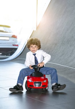 Kravat takan ve gömlek giyen sevimli, komik bir çocuk küçük kırmızı bir oyuncak arabanın üzerinde oturuyor. Kıvırcık saçlı esmer çocuk oto kuaförde eğleniyor. İş galerisi kavramı