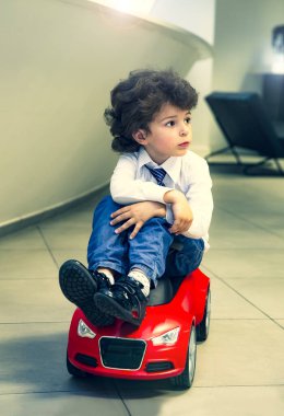 Kravat takan ve gömlek giyen sevimli, komik bir çocuk küçük kırmızı bir oyuncak arabanın üzerinde oturuyor. Kıvırcık saçlı esmer çocuk oto kuaförde eğleniyor. İş galerisi kavramı