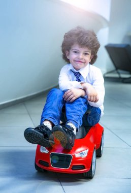 Kravat takan ve gömlek giyen sevimli, komik bir çocuk küçük kırmızı bir oyuncak arabanın üzerinde oturuyor. Kıvırcık saçlı esmer çocuk oto kuaförde eğleniyor. İş galerisi kavramı
