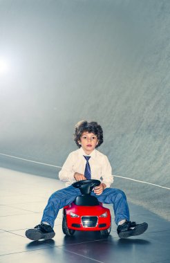 Kravat takan ve gömlek giyen sevimli, komik bir çocuk küçük kırmızı bir oyuncak arabanın üzerinde oturuyor. Kıvırcık saçlı esmer çocuk oto kuaförde eğleniyor. İş galerisi kavramı