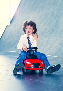 Kravat takan ve gömlek giyen sevimli, komik bir çocuk küçük kırmızı bir oyuncak arabanın üzerinde oturuyor. Kıvırcık saçlı esmer çocuk oto kuaförde eğleniyor. İş galerisi kavramı