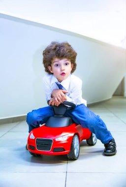 Kravat takan ve gömlek giyen sevimli, komik bir çocuk küçük kırmızı bir oyuncak arabanın üzerinde oturuyor. Kıvırcık saçlı esmer çocuk oto kuaförde eğleniyor. İş galerisi kavramı
