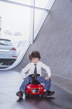 Kravat takan ve gömlek giyen sevimli, komik bir çocuk küçük kırmızı bir oyuncak arabanın üzerinde oturuyor. Kıvırcık saçlı esmer çocuk oto kuaförde eğleniyor. İş galerisi kavramı