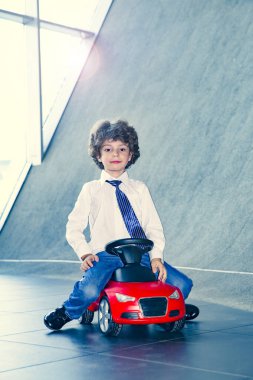Kravat takan ve gömlek giyen sevimli, komik bir çocuk küçük kırmızı bir oyuncak arabanın üzerinde oturuyor. Kıvırcık saçlı esmer çocuk oto kuaförde eğleniyor. İş galerisi kavramı