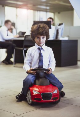 Kravat takan ve gömlek giyen sevimli, komik bir çocuk küçük kırmızı bir oyuncak arabanın üzerinde oturuyor. Kıvırcık saçlı esmer çocuk oto kuaförde eğleniyor. İş galerisi kavramı