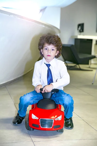 Kravat takan ve gömlek giyen sevimli, komik bir çocuk küçük kırmızı bir oyuncak arabanın üzerinde oturuyor. Kıvırcık saçlı esmer çocuk oto kuaförde eğleniyor. İş galerisi kavramı