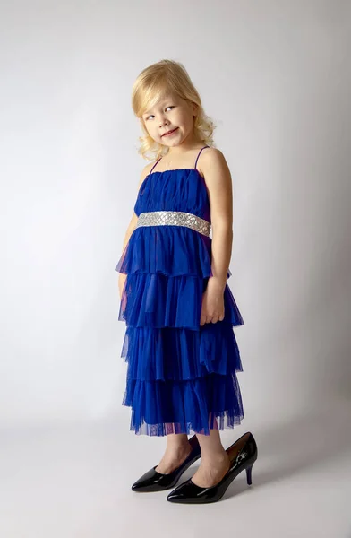 Little princess girl in fashion blue dress wearing big mothers patent heels shoes looks suspiciously and incredulously to the side on gray background. Free space for text mockup