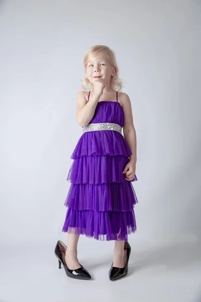 Little princess excited girl in fashion purple dress wearing big mothers sparkle heels shoes on white background. Free space for text mockup