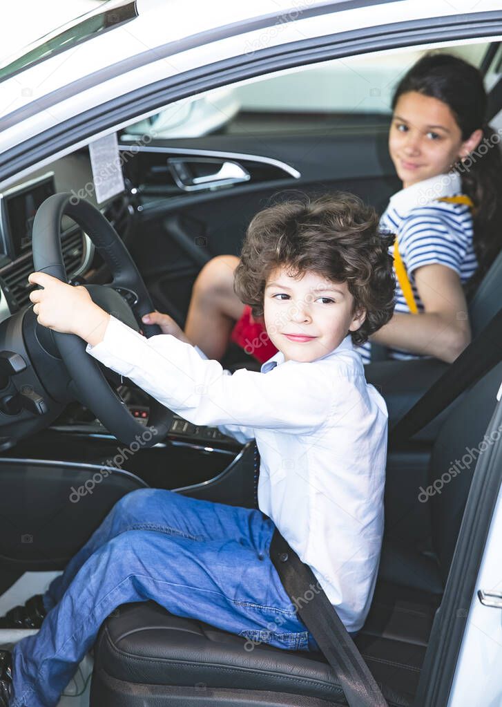 Children observing and testing new car in dealership. Little boy sitting in driver's seat, car cabin. Little boy holding hands on steering wheel, smiling.