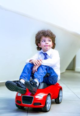 Kravat takan ve gömlek giyen sevimli, komik bir çocuk küçük kırmızı bir oyuncak arabanın üzerinde oturuyor. Kıvırcık saçlı esmer çocuk oto kuaförde eğleniyor. İş galerisi kavramı