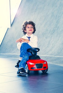 Kravat takan ve gömlek giyen sevimli, komik bir çocuk küçük kırmızı bir oyuncak arabanın üzerinde oturuyor. Kıvırcık saçlı esmer çocuk oto kuaförde eğleniyor. İş galerisi kavramı