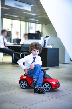 Kravat takan ve gömlek giyen sevimli, komik bir çocuk küçük kırmızı bir oyuncak arabanın üzerinde oturuyor. Kıvırcık saçlı esmer çocuk oto kuaförde eğleniyor. İş galerisi konsepti. Seçici odak.