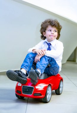 Kravat takan ve gömlek giyen sevimli, komik bir çocuk küçük kırmızı bir oyuncak arabanın üzerinde oturuyor. Kıvırcık saçlı esmer çocuk oto kuaförde eğleniyor. İş galerisi kavramı