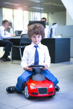 Kravat takan ve gömlek giyen sevimli, komik bir çocuk küçük kırmızı bir oyuncak arabanın üzerinde oturuyor. Kıvırcık saçlı esmer çocuk oto kuaförde eğleniyor. İş galerisi kavramı