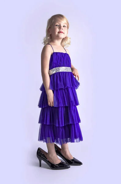 Little princess excited girl in fashion lilac dress wearing big mothers sparkle heels shoes on light background. Free space for text mockup