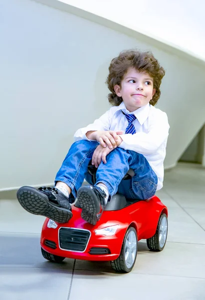 Kravat takan ve gömlek giyen sevimli, komik bir çocuk küçük kırmızı bir oyuncak arabanın üzerinde oturuyor. Kıvırcık saçlı esmer çocuk oto kuaförde eğleniyor. İş galerisi kavramı