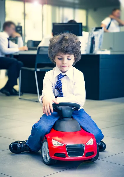 Kravat takan ve gömlek giyen sevimli, komik bir çocuk küçük kırmızı bir oyuncak arabanın üzerinde oturuyor. Kıvırcık saçlı esmer çocuk oto kuaförde eğleniyor. İş galerisi kavramı