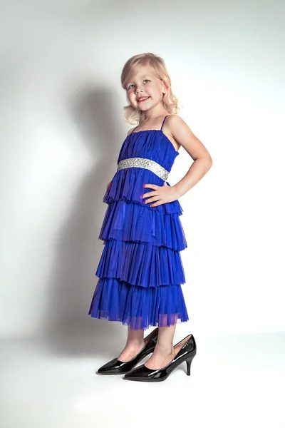 Little princess excited girl in fashion blue dress wearing big mothers sparkle heels shoes on white background. Free space for text mockup