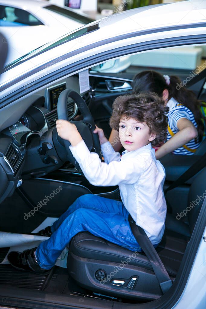 Children observing and testing new car in dealership. Little boy sitting in driver's seat, car cabin. Little boy holding hands on steering wheel, smiling. Selective focus. Light blur.