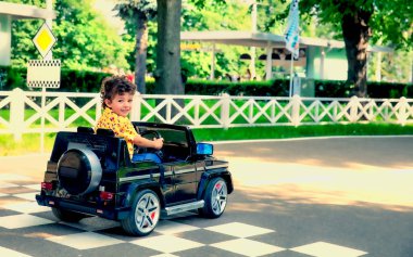 Sarı tişörtlü 4 yaşındaki tatlı çocuk parkta siyah elektrikli bir araba sürüyor.