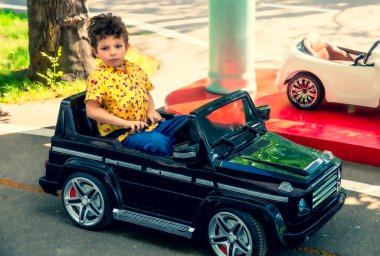 Sarı tişörtlü sevimli 4 yaşındaki çocuk parkta siyah elektrikli bir araba sürüyor. Çocuk oyuncak bir arabaya bağlanır.