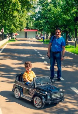 Sarı tişörtlü sevimli 4 yaşındaki çocuk parkta siyah elektrikli bir araba sürüyor. Ailesi yazın lunaparkta.
