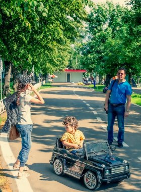Sarı tişörtlü sevimli 4 yaşındaki çocuk parkta siyah elektrikli bir araba sürüyor. Ailesi yazın lunaparkta.