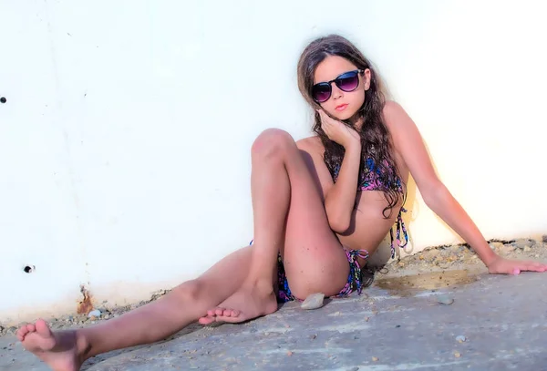 Mayo giymiş, güneş gözlüğü takmış, duvara poz veren ince bir kızın portresi.