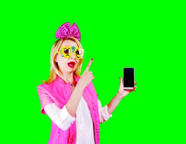 Karnaval güneş gözlüklü ve pembe elbiseli neşeli bir kadın telefon ekranı gösteriyor. Parlak renkli arka planda izole edilmiş bir fotokopi alanını parmağıyla işaret ediyor. Kameraya bakıyorum.