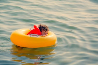 Büyük sarı bir yüzme çemberinde oturan can yeleği giymiş küçük mutlu çocuk denizde yüzüyor. Boşluğu kopyala