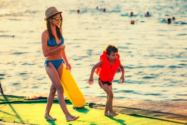 Şişme yelek giyen küçük kardeş ve denizin arka planında beton bir iskelede yürüyen şişme halkalı ablası..