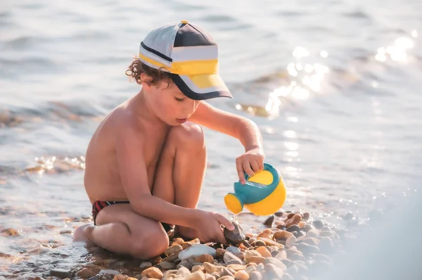 小男孩在卵石沙滩上 一个小男孩坐在海滩上玩水壶 — 图库照片