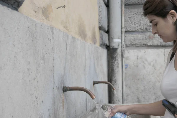 A woman Filling a bottle with drinkable water in a public fountain, Rome, Italy