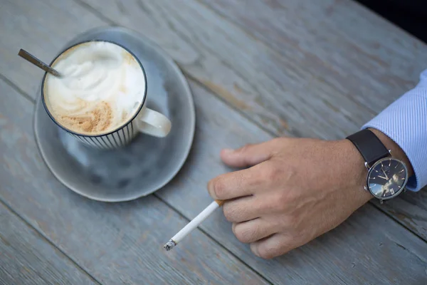 Closeup shot of a cup of coffee and a mans hand with a cigarette daytime