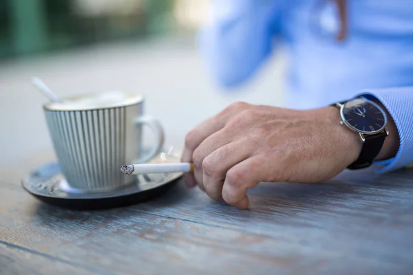 Closeup shot of a cup of coffee and a mans hand with a cigarette daytime