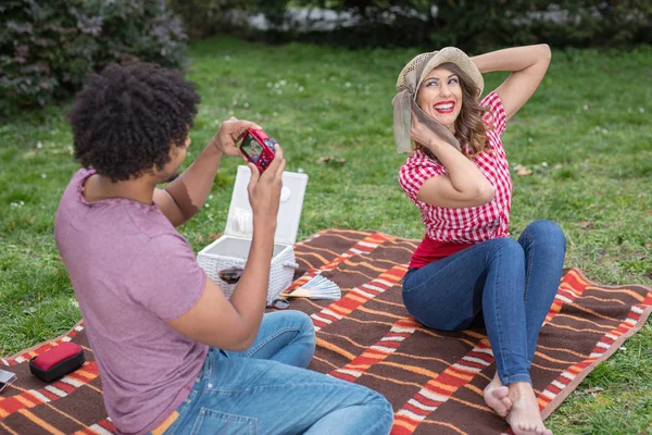 Beautiful young couple taking pictures with a camera of each other outside on a picnic blanket while smiling