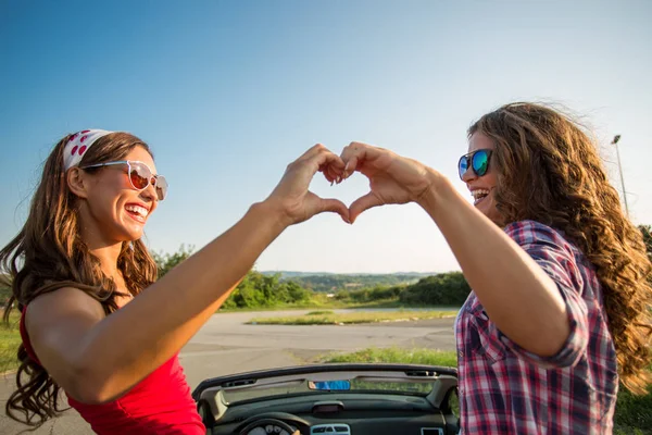 Two beautiful young women making a heart with their hands in a car while smiling at each other