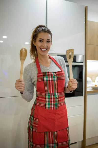 Beautiful young woman standing in a kitchen wearing an apron while holding wooden spoons and smiling