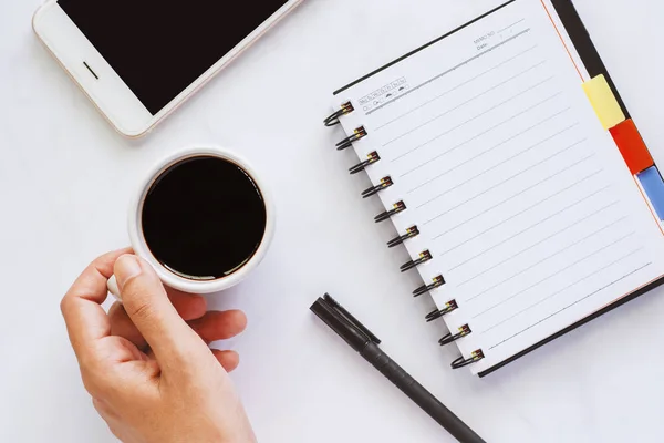Kahve spiral defter, kalem ve beyaz mermer arka planda iş kavramı, dinlenmek için smartphone ile tutan el