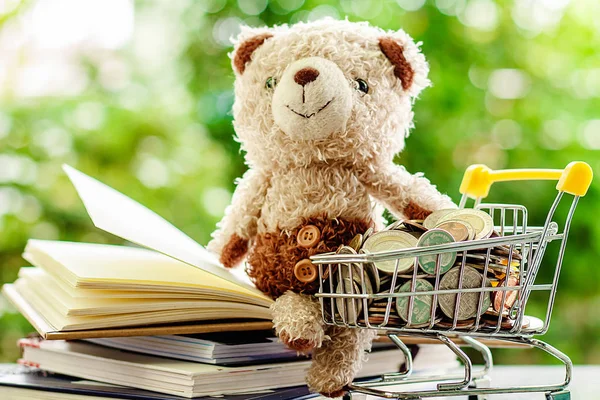 Smiling teddy bear doll sitting on stack of opened book with full of money coin in mini shopping cart or trolley against blurred natural green background for save money and education financial concept