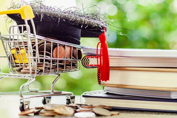 Square academic cap on money coin in mini shopping cart or trolley and stack of books against blurred natural green background for save money and education financial concept