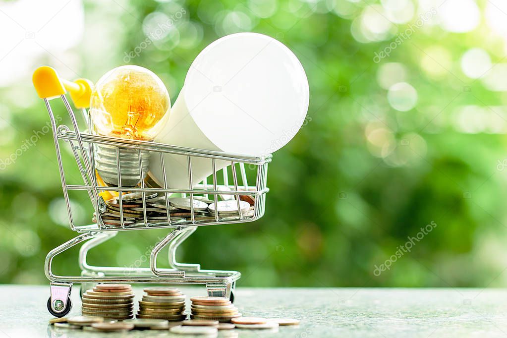 Led lamp and glowing light bulb in mini shopping cart or trolley  with money coins against blurred natural green background for finance, saving energy and environment concept